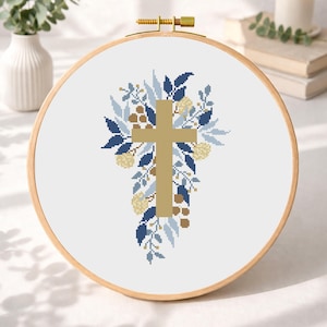 May include: Cross-stitch embroidery in a wooden hoop. The design features a gold-coloured cross surrounded by blue, light blue, and tan floral elements. The background is white, and the hoop is made of light-coloured wood.