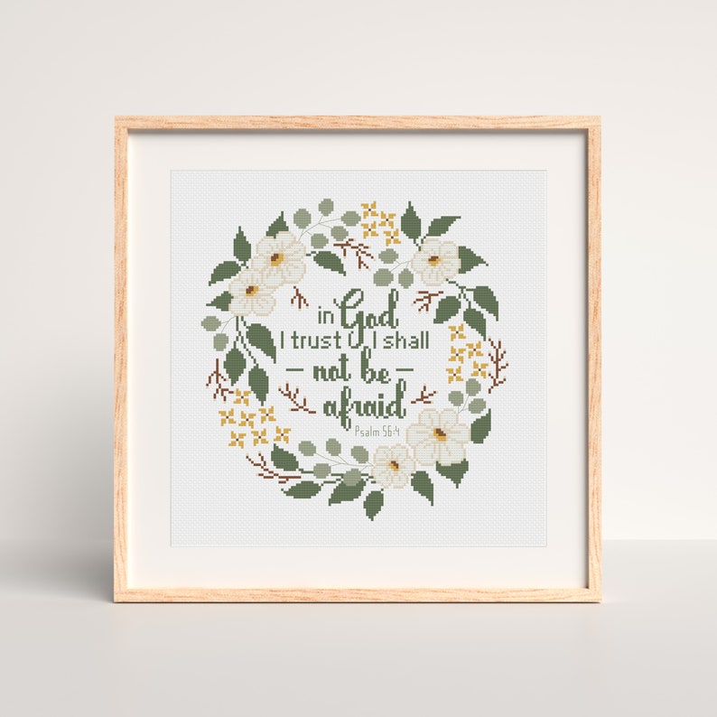 May include: A framed cross-stitch artwork featuring a floral wreath design. The wreath surrounds the text "in God I trust I shall not be afraid" in a script font. The wreath includes white flowers, green leaves, and yellow accents. The frame is made of light-colored wood.