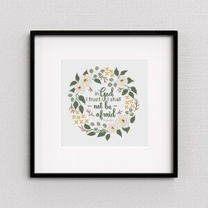 May include: A framed cross-stitch artwork featuring a floral wreath design. The wreath is composed of white flowers, green leaves, and yellow accents, surrounding the text "in God I trust I shall not be afraid" from Psalm 56:4. The frame is black.