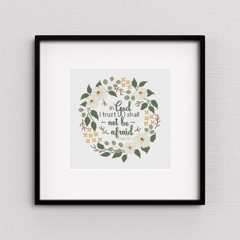 May include: A framed cross-stitch artwork featuring a floral wreath design. The wreath is composed of white flowers, green leaves, and yellow accents, surrounding the text "in God I trust I shall not be afraid" from Psalm 56:4. The frame is black.