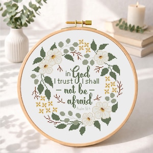 May include: A cross-stitch embroidery hoop featuring the quote "in God I trust, I shall not be afraid" surrounded by a floral wreath. The wreath includes white flowers, green leaves, and yellow accents. The text is in a dark green color.