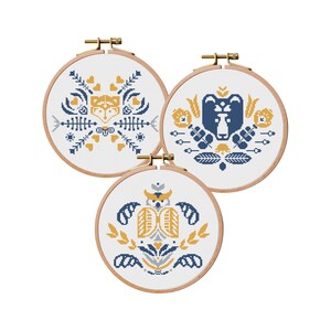 May include: Three embroidery hoops with white fabric and colorful cross-stitch designs. The designs feature a fox, a bear, and an owl, all with a geometric style and a color palette of blue, yellow, and gray.