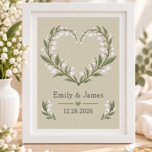 May include: A framed cross-stitch pattern on a light beige background featuring a heart-shaped wreath of white lily of the valley flowers and green leaves. The text reads Emily & James above a small heart and the date 12.26.2026 in dark gray.
