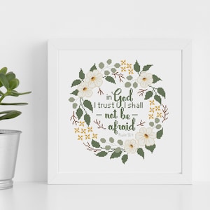May include: A framed cross-stitch artwork featuring a floral wreath design. The wreath surrounds the text "in God I trust I shall not be afraid" in green lettering. White flowers and green leaves are incorporated into the design.