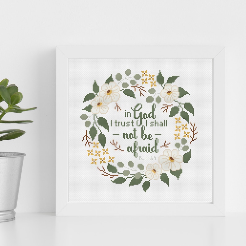 May include: A framed cross-stitch artwork featuring a floral wreath design. The wreath surrounds the text "in God I trust I shall not be afraid" in green lettering. White flowers and green leaves are incorporated into the design.