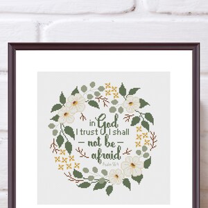 May include: A framed cross-stitch artwork featuring a floral wreath design. The wreath surrounds the text "in God I trust I shall - not be - afraid" from Psalm 56:4. The design includes white flowers, green leaves, and yellow accents.