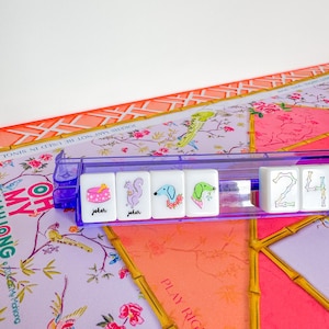 Op de afbeelding: Een close-up van een Mahjong-set met witte tegels met kleurrijke illustraties en het woord "joker". De tegels worden vastgehouden in een doorzichtig paars rek, geplaatst op een spelbord met bloemenpatroon met de tekst "PLAY RIGHT".