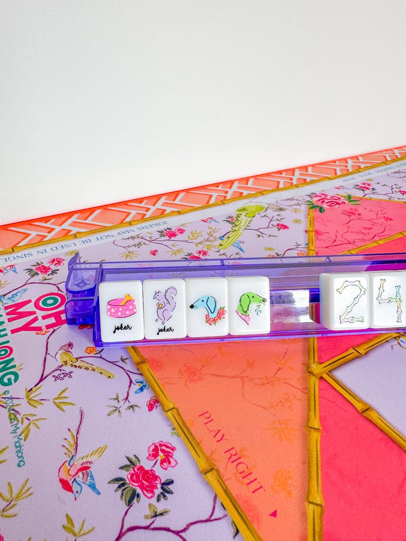 Op de afbeelding: Een close-up van een Mahjong-set met witte tegels met kleurrijke illustraties en het woord "joker". De tegels worden vastgehouden in een doorzichtig paars rek, geplaatst op een spelbord met bloemenpatroon met de tekst "PLAY RIGHT".