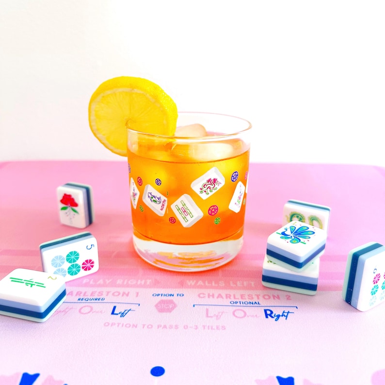 May include: A cocktail in a clear glass with ice and a lemon slice, surrounded by Mahjong tiles on a pink surface. The glass is decorated with Mahjong tile designs. The tiles are white, blue, and green.
