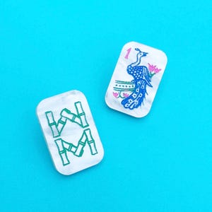 Puede incluir: Dos fichas de Mahjong blancas con diseños verdes y azules sobre un fondo azul brillante. Una ficha presenta un pavo real con una flor rosa y el número 1. La otra ficha tiene un diseño de bambú verde.