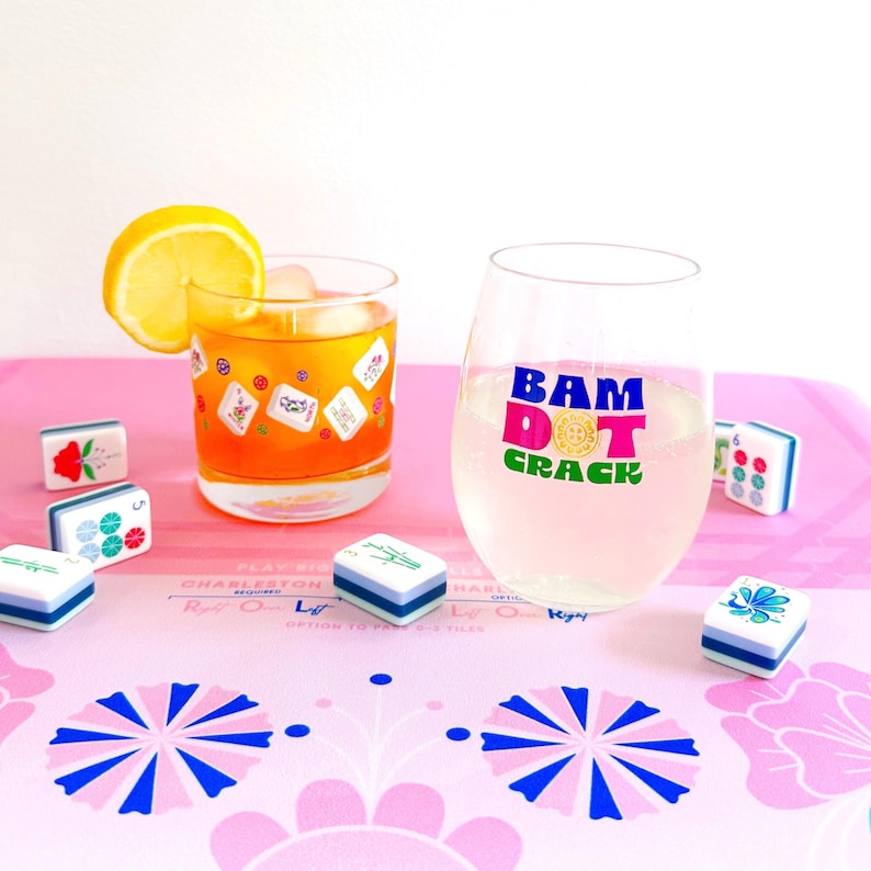 May include: A glass of orange liquid with ice and a lemon slice, and a stemless wine glass with the text "BAM DOT CRACK". Several Mahjong tiles are scattered on a pink surface with a floral design.