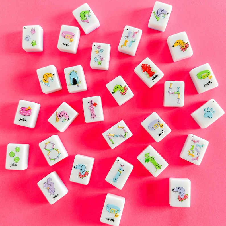 Op de afbeelding: Witte mahjongtegels met kleurrijke illustraties met honden als thema en tekst. De tegels tonen verschillende hondenrassen, pootafdrukken en woorden als "oost" en "noord". De achtergrond is felroze.