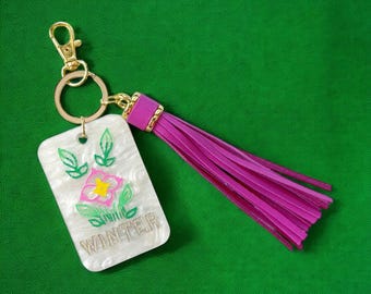 May include: A rectangular keychain with a white, marbled design, featuring a pink and yellow flower with green leaves, and the word "WINTER" in gold lettering. It has a gold-colored clasp and key ring, and a long, magenta tassel.