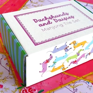 Op de afbeelding: Een decoratieve doos voor een Mahjong-tegelset. De doos is wit met een paars omkaderd label met de tekst "Dachshunds and Daisies". De doos heeft een kleurrijk patroon van teckels en madeliefjes. De zijkant van de doos is groen en blauw gestreept.