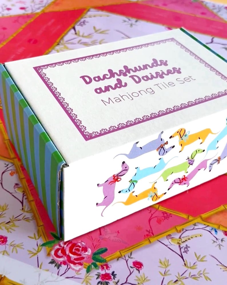 Op de afbeelding: Een decoratieve doos voor een Mahjong-tegelset. De doos is wit met een paars omkaderd label met de tekst "Dachshunds and Daisies". De doos heeft een kleurrijk patroon van teckels en madeliefjes. De zijkant van de doos is groen en blauw gestreept.
