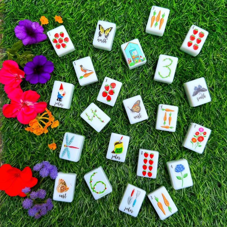 Pode incluir: Um conjunto de pe&ccedil;as retangulares brancas de mahjong com ilustra&ccedil;&otilde;es coloridas de elementos de jardim, incluindo morangos, cenouras, borboletas, flores, um gnomo de jardim, uma estufa e ferramentas, com etiquetas north, south, east, west e joker.