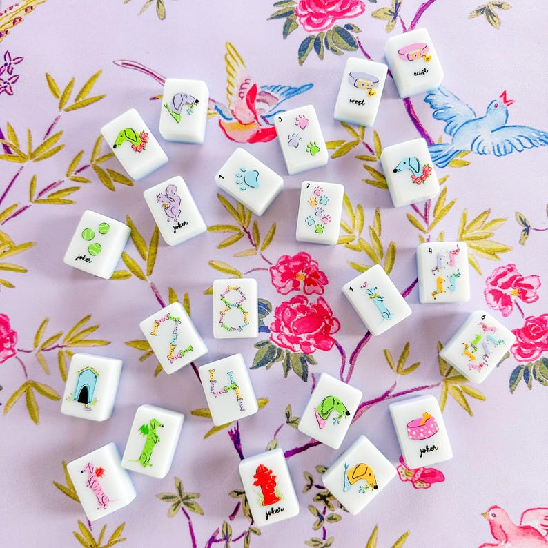 Op de afbeelding: Een verzameling witte Mahjong-tegels met kleurrijke illustraties van honden, eekhoorns en andere ontwerpen. De tegels zijn verspreid op een bloemmotief achtergrond, sommige met het label "joker".