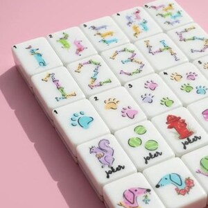 Op de afbeelding: Een set witte Mahjong-tegels met kleurrijke illustraties. De tegels tonen cijfers, pootafdrukken, hondenillustraties en het woord "joker". De achtergrond is effen roze.