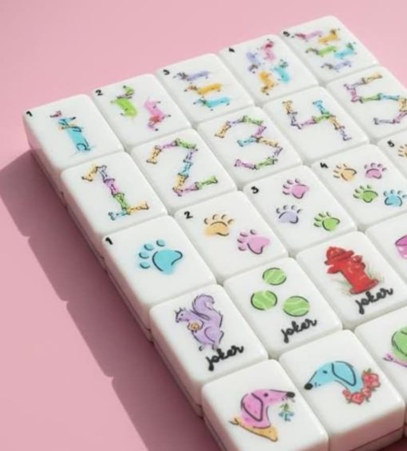 Op de afbeelding: Een set witte Mahjong-tegels met kleurrijke illustraties. De tegels tonen cijfers, pootafdrukken, hondenillustraties en het woord "joker". De achtergrond is effen roze.