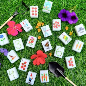 Pode incluir: Um conjunto de pe&ccedil;as retangulares brancas de mahjong com ilustra&ccedil;&otilde;es coloridas de artigos de jardim, n&uacute;meros e insetos, espalhadas entre flores vermelhas e roxas, um ancinho de jardim e uma pequena p&aacute; de metal preto.