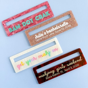 May include: Four rectangular acrylic name tags with text. One is pink with teal text reading "BAM DOT CRAK". Another is glittery with "Julie's bachelorette Table Rock Lake June 10, 2026". A third is white with rainbow text "get your mahj on". The last is brown with "mahjong girls weekend Boca Raton, FL April 3, 2026".