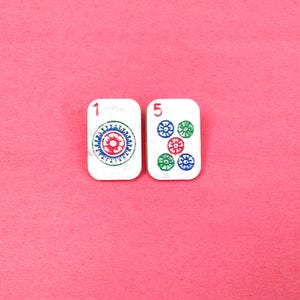 May include: Two white Mahjong tiles with red, blue, and green designs on a pink background. One tile shows the number 1 and a circular pattern. The other tile shows the number 5 and five circular patterns.