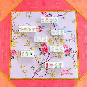Op de afbeelding: Een decoratieve Mahjong-set met een roze en gouden bamboe-stijl frame. Het speeloppervlak heeft een bloemen- en vogelontwerp in roze, groen en wit. De tegels zijn wit met kleurrijke illustraties en cijfers.