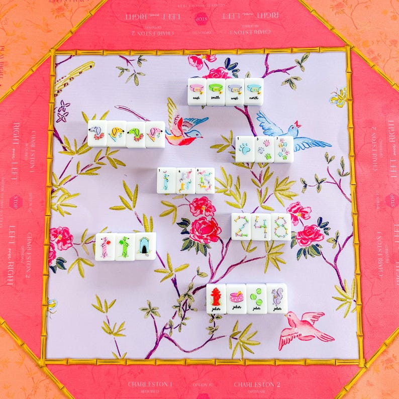 Op de afbeelding: Een decoratieve Mahjong-set met een roze en gouden bamboe-stijl frame. Het speeloppervlak heeft een bloemen- en vogelontwerp in roze, groen en wit. De tegels zijn wit met kleurrijke illustraties en cijfers.