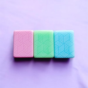 Op de afbeelding: Drie rechthoekige zepen in pastelkleuren: roze, groen en blauw. Elke zeep heeft een geometrisch kubuspatroon. De zepen zijn in een rij gerangschikt tegen een paarse achtergrond.