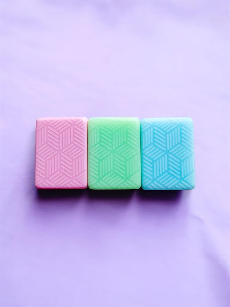 Op de afbeelding: Drie rechthoekige zepen in pastelkleuren: roze, groen en blauw. Elke zeep heeft een geometrisch kubuspatroon. De zepen zijn in een rij gerangschikt tegen een paarse achtergrond.