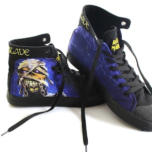 Può includere: Un paio di sneakers alte con lacci e suole neri. Le tomaie in tela presentano un motivo a fulmini blu e una grafica di una mummia. La parola "SLAVE" è stampata in oro sul colletto e "IRON MAIDEN" sul lato.