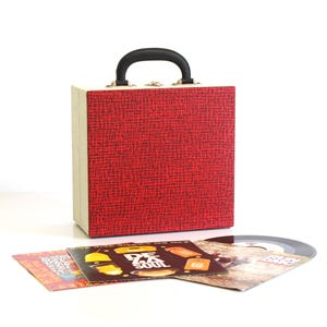 Puede incluir: Un estuche rojo para discos de vinilo con asa negra y laterales color crema. Varias portadas de discos de vinilo y un disco de 45 rpm están frente al estuche. Las portadas tienen gráficos y texto coloridos.