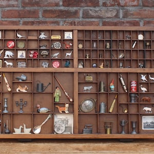 Puede incluir: Una bandeja de imprenta rectangular de madera con numerosos compartimentos pequeños, cada uno con una variedad de figuras en miniatura, objetos metálicos pequeños, azulejos de cerámica diminutos y baratijas, incluyendo una lata roja.