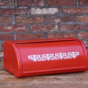 Peut inclure: Un coffre à pain en métal rouge avec un dessus incurvé et un motif floral blanc. Le coffre à pain est rectangulaire et repose sur une surface en bois devant un mur de briques. Un article de rangement de cuisine vintage.