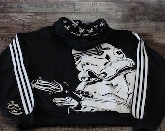 May include: Black hoodie featuring a large white graphic of a Stormtrooper with a blaster. The hoodie has white stripes down the sleeves and a small Stormtrooper logo on the cuff. The hood lining has a white pattern.