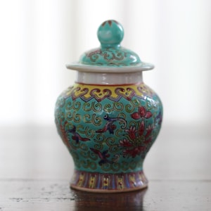 Op de afbeelding: Een kleine, tealblauwe porseleinen pot met een deksel. De pot is versierd met een bloemenpatroon in tinten geel, rood en groen.