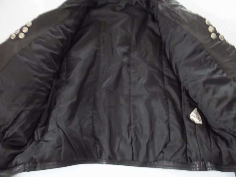 May include: Black leather jacket with silver-tone stud embellishments on the shoulders. The interior is lined with black satin fabric. The jacket is open, revealing the inside lining and construction.