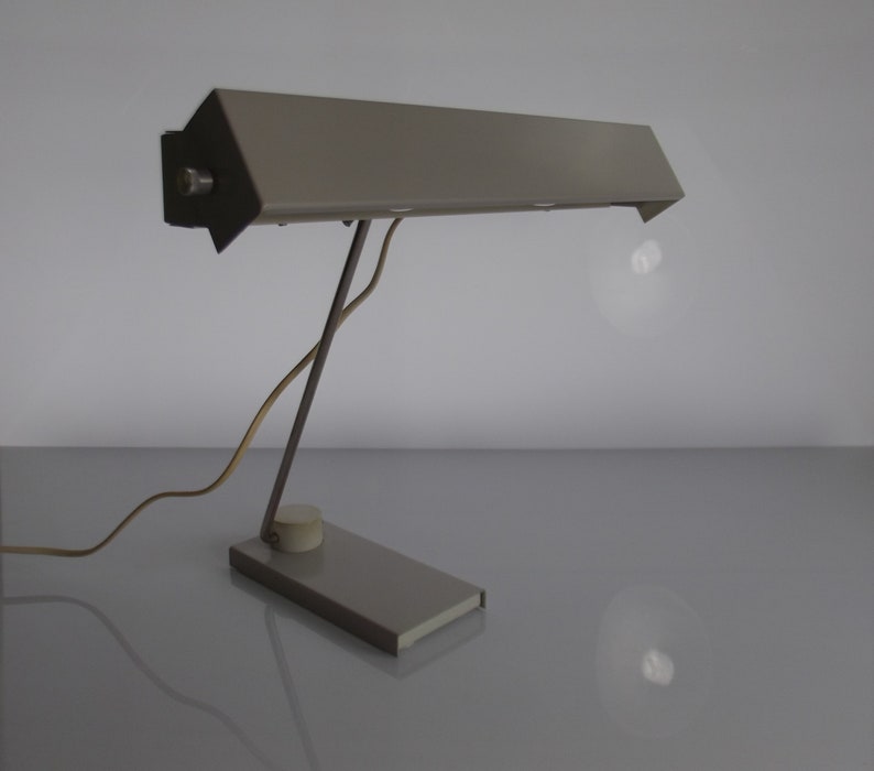 K&ouml;nnte beinhalten: Eine graue Metall-Tischleuchte mit einem langen, rechteckigen Schirm und einem flexiblen Arm. Die Lampe hat ein schlichtes, modernes Design.
