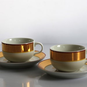 Op de afbeelding: Twee witte theekopjes met gouden en rode versieringen en bijpassende schoteltjes. De kopjes en schoteltjes hebben een gouden band rond de rand.