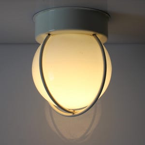 May include: A white ceiling light with a spherical, opaque glass shade encased in a metal cage. The light fitting has a white metal base and is mounted on a white ceiling. It emits a warm, inviting light.