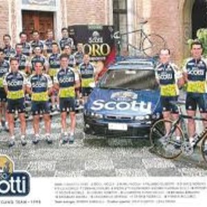 May include: A group of cyclists in matching yellow and blue jerseys stand in front of a white car with the word "Scotti" on the side. The cyclists are holding bicycles and are smiling. The car is a Fiat.