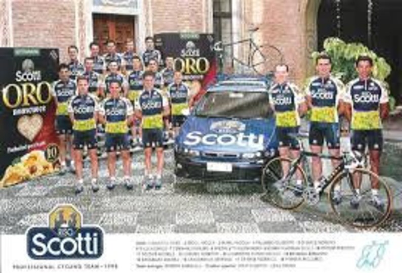 May include: A group of cyclists in matching yellow and blue jerseys stand in front of a white car with the word "Scotti" on the side. The cyclists are holding bicycles and are smiling. The car is a Fiat.