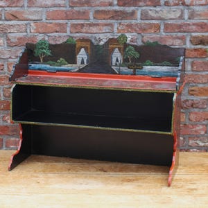 May include: A wooden shelf with a painted landscape scene on the top. The shelf is painted black with red accents and has two shelves. The landscape features trees, water, and buildings.
