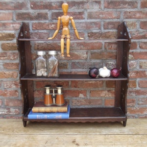 Può includere: Una mensola in legno scuro a tre livelli con bordi decorativi. I ripiani contengono barattoli di vetro, libri, binocoli, cipolle e un manichino di artista in legno. Lo sfondo è un muro di mattoni rossi.