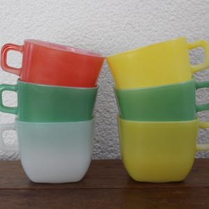 May include: A stack of six colorful vintage coffee mugs. The mugs are in shades of red, green, yellow, and white. Each mug has a square handle. The mugs are stacked on a wooden surface.