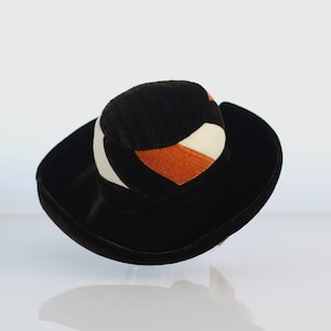 Op de afbeelding: Een zwarte corduroy hoed met een brede rand en een geometrisch patroon van witte en oranje stof.