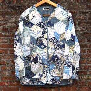 Op de afbeelding: Een gewatteerde jas met een patchwork-ontwerp in tinten blauw, wit en crème, hangend aan een houten hanger tegen een bakstenen muur. De jas heeft een V-hals, knoopsluiting en twee voorzakken. Het patchwork bevat bloemen- en geometrische patronen.