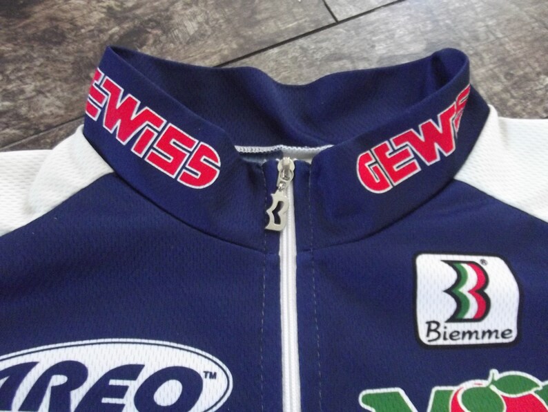 May include: A blue and white cycling jersey with red "GEWISS" text on the collar, a white zipper, and a white "Biemme" logo with a green, white, and red stripe design.