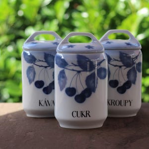 Op de afbeelding: Drie witte keramische potten met blauwe en witte kersenmotieven. De potten zijn gelabeld met "KAVA", "CUKR" en "KROUPY".