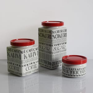 Könnte beinhalten: Drei weiße Keramikdosen mit leuchtend roten Deckeln, jeweils in quadratischer oder zylindrischer Form, bedruckt mit schwarzer Typografie in verschiedenen Sprachen. Die Dosen zeigen Wörter wie Kaffee, Zucker und Tee in diversen Schriftarten.